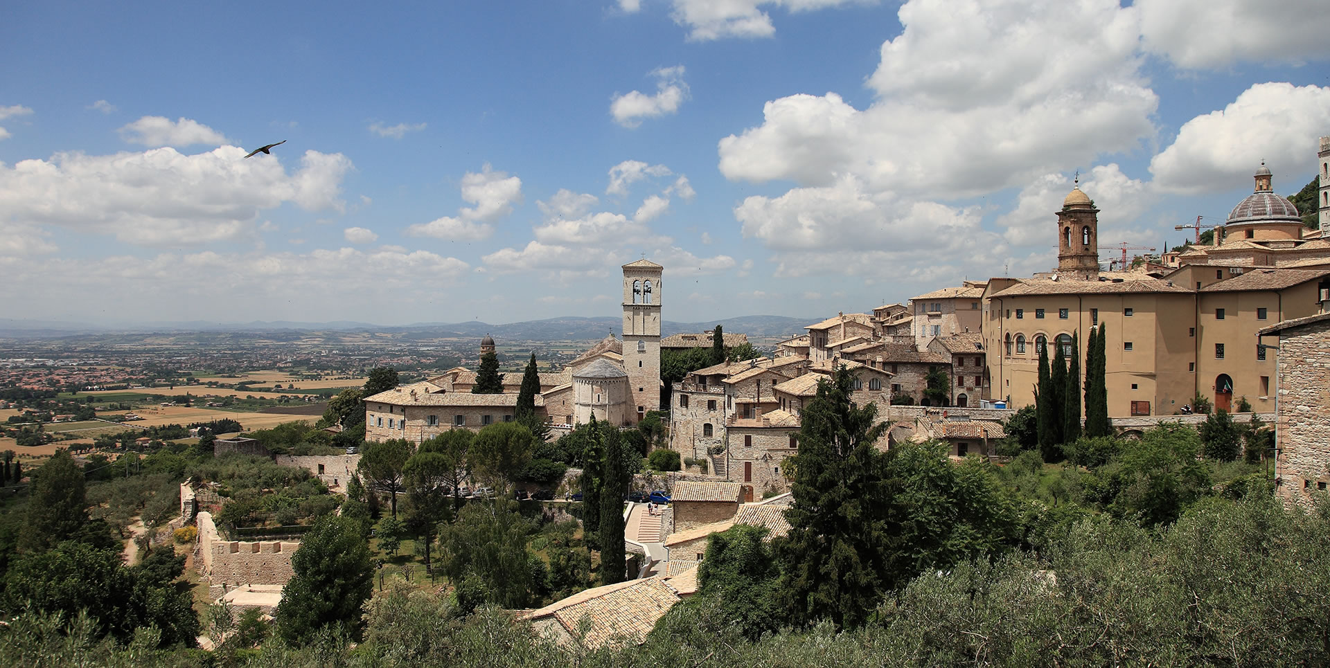 Panorama di Assisi con edifici storici in pietra, campanili e torri sotto un cielo sereno ma con qualche nuvola